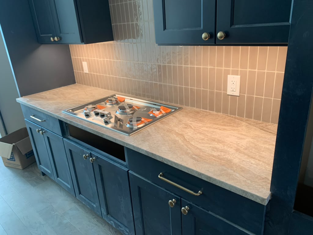 Beige stone countertop with natural veining and a built-in cooktop cutout, navy blue cabinets and tile backsplash